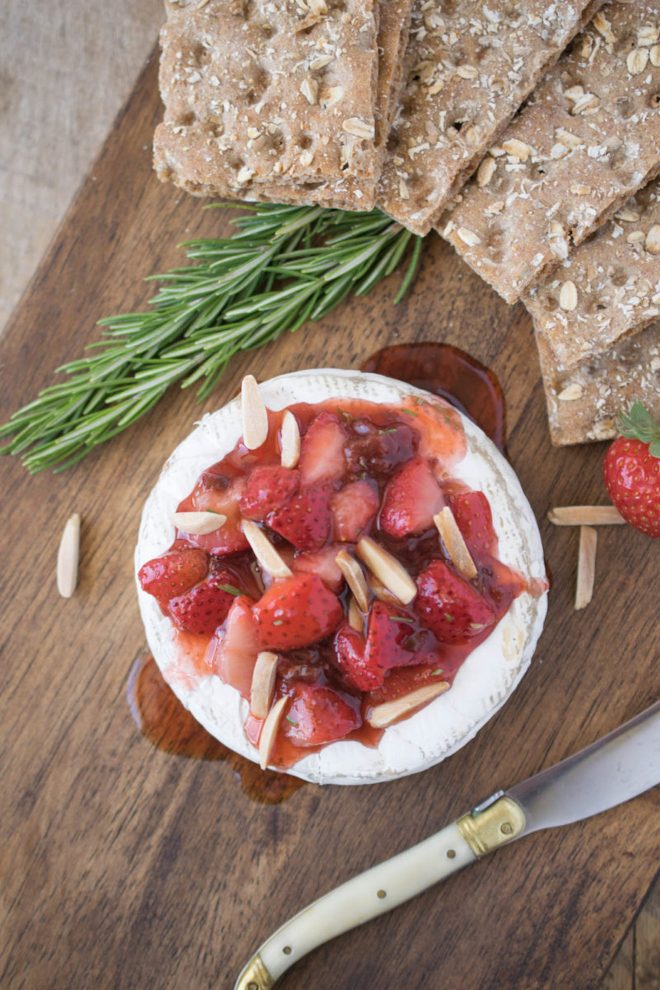 A wheel of brie topped with strawberries, fresh rosemary and almonds on a board with a knife and crackers