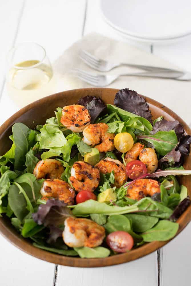 A wood bowl full of lettuce topped with grilled shrimp, red and yellow tomatoes with forks and a glass of wine
