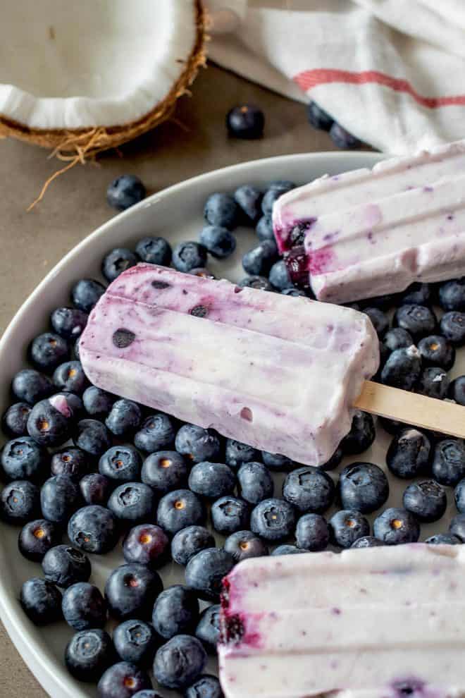 A closeup of a blueberry coconut frozen yogurt popsicle on top of fresh blueberries