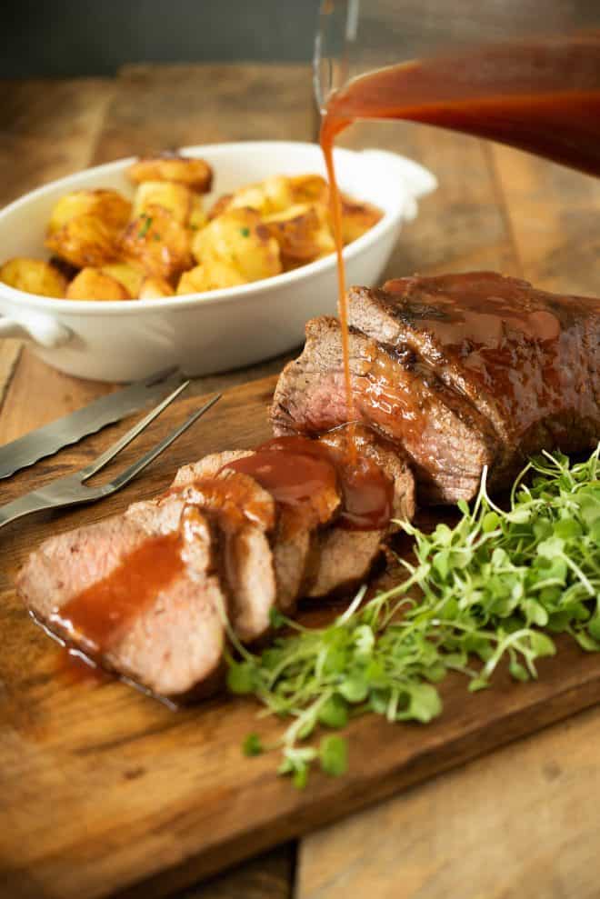 Drizzling barbecue sauce over sliced Tri tip with roast potatoes in the background