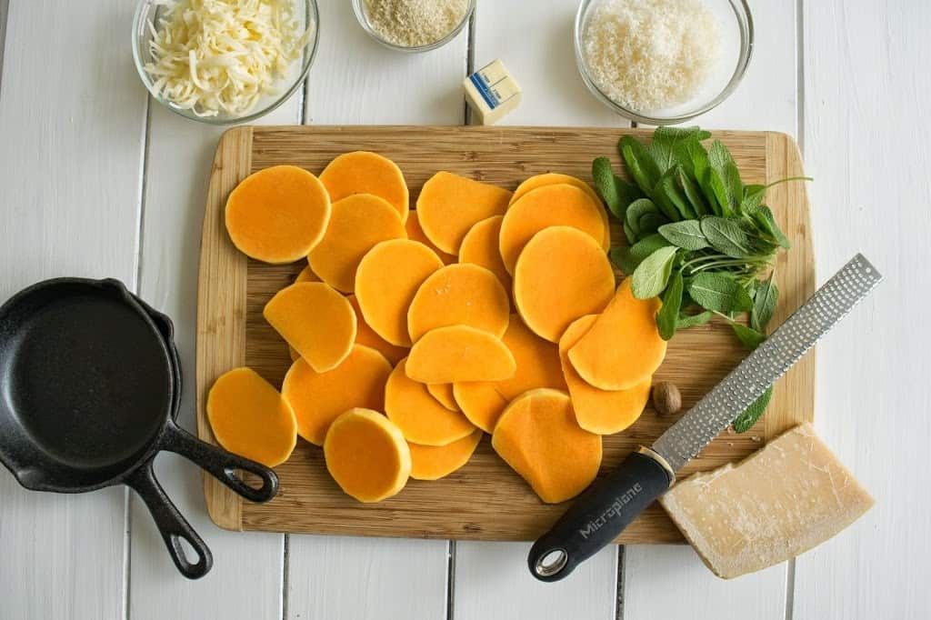A cutting board with sliced butternut squash, fresh sage, parmesan cheese and grated fontina viewed from overhead
