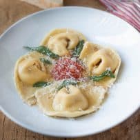 A round white pasta bowl with 4 butternut squash filled cappellacc with a small amount of marinara sauce in the center with crispy sage leaves and grated Parmesan cheesei