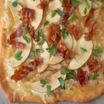 A closeup of the topping on a flatbread pizza of fontina cheese, apple, caramelized onions, bacon pieces and green parsley