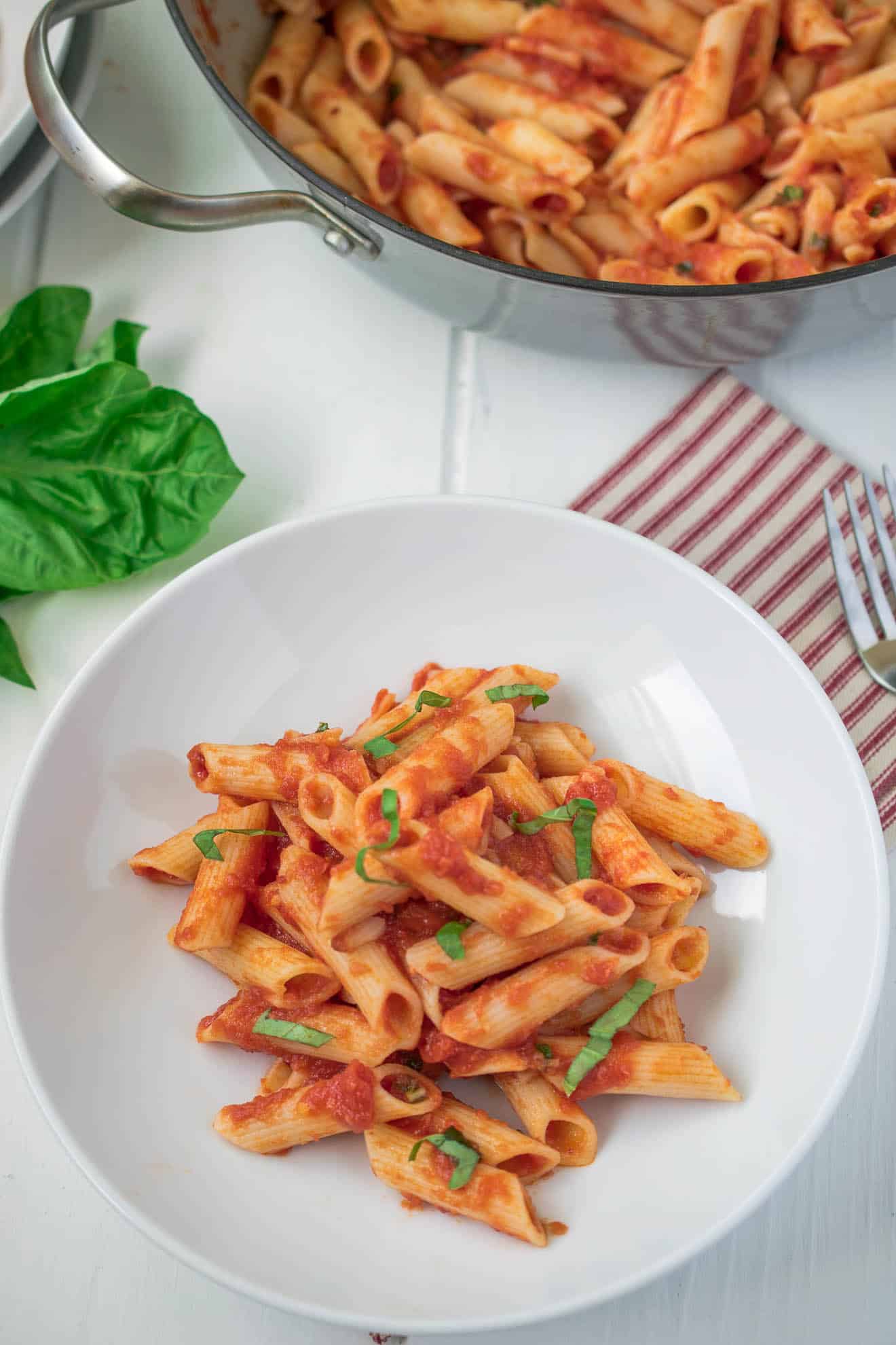 A bowl of classic penne all'arrabbiata with fresh basil and a pan it was cooked in