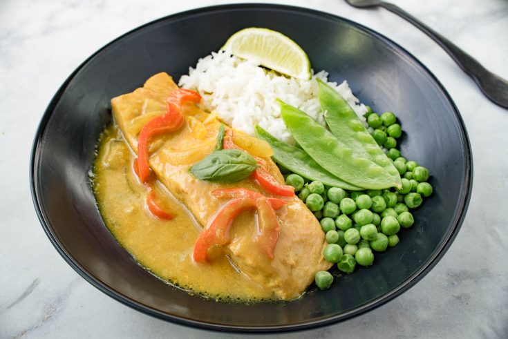 A filet of salmon bathed in coconut curry sauce with fresh peas, snow peas and white rice in a black bowl