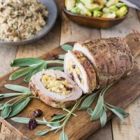 Apple cranberry cornbread stuffed pork loin served on a cutting board garnished with fresh sage leaves with a bowl of Brussels sprouts and rice in background