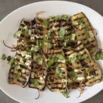 Slices of aubergine/eggplant with grill marks on a plate topped with curry yogurt sauce, crispy onions, raisins and coriander/cilantro.
