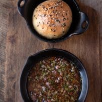 A small cast iron bowl that holds a crusty bread roll alongside a cast iron skillet with bacon, sage butter