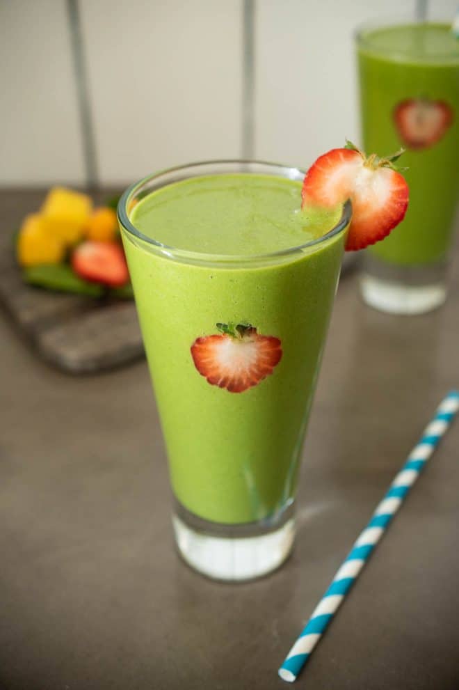 A glass of detox green smoothie with a straw and fresh strawberry slices