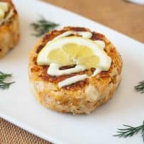 A white serving plate with crab cakes topped with lemon scallion sauce, lemon and fresh dill