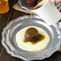 Pouring toffee sauce over sticky toffee pudding on a pewter plate with custard