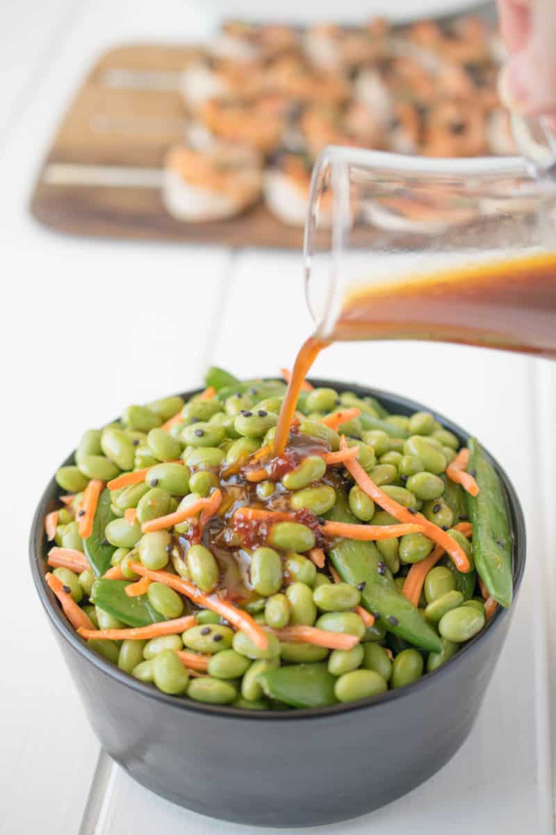 Pouring sesame ginger dressing onto a bowl of edamame and snap peas