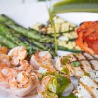 Pouring herb sauce over grilled shrimp, chicken and vegetables on a white platter