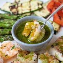 Herb sauce in a grey bowl with a shrimp on a fork dipping it into the sauce surrounded by more shrimp and grilled vegetables