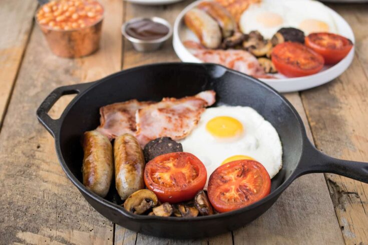 Fried egg, bacon, sausage, tomato, mushrooms in a cast iron pan.