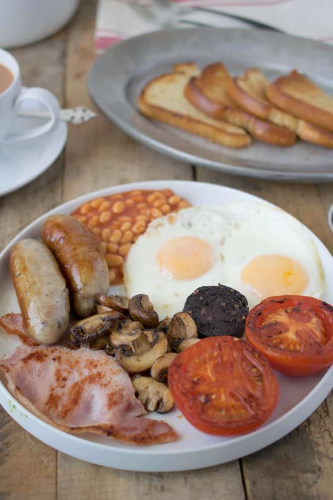 A large white plate with fried eggs, baked beans, sausages, mushrooms, bacon, tomatoes and black pudding.