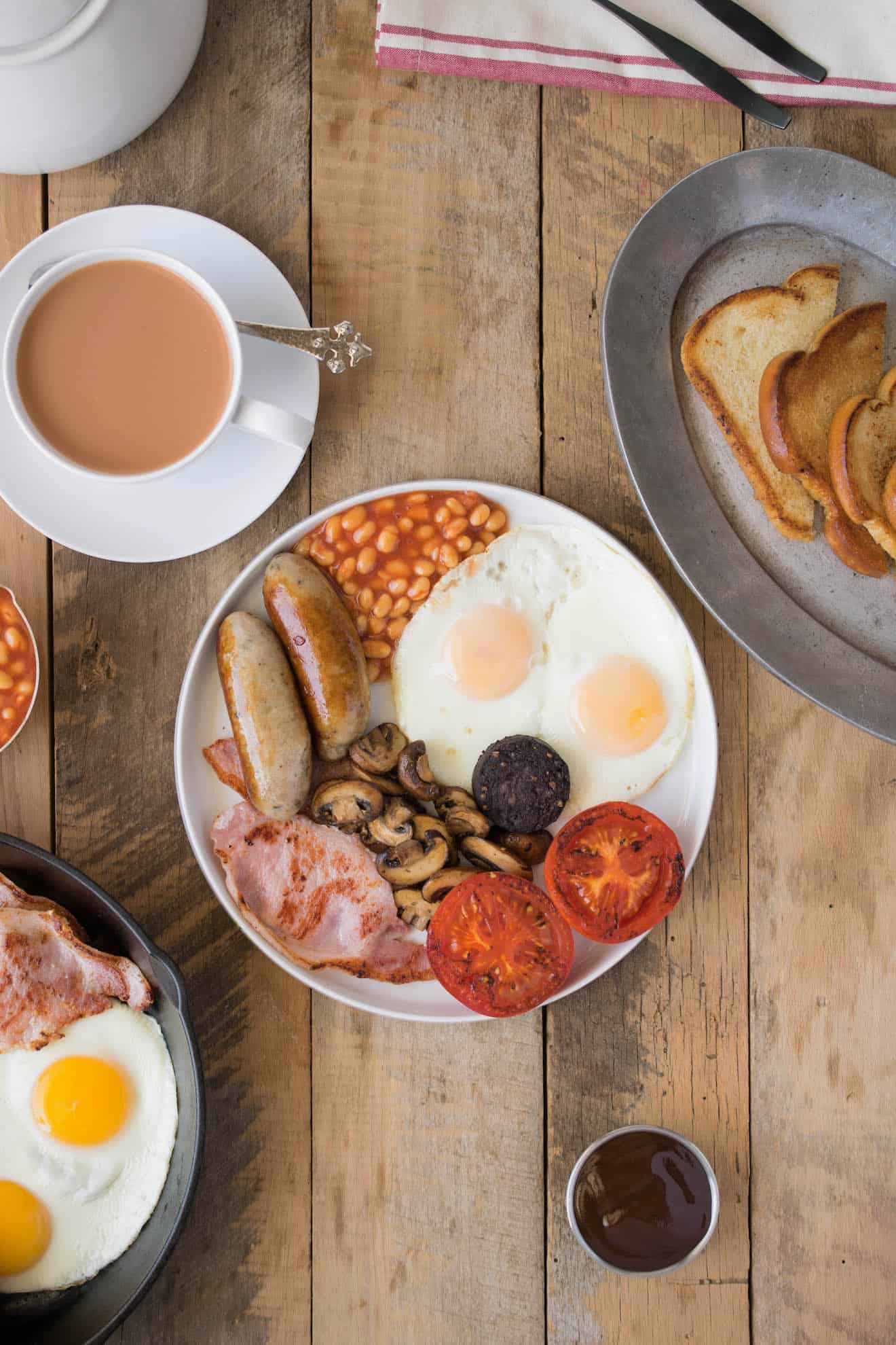 A Full English breakfast on a white plate with fried break and a cup of tea
