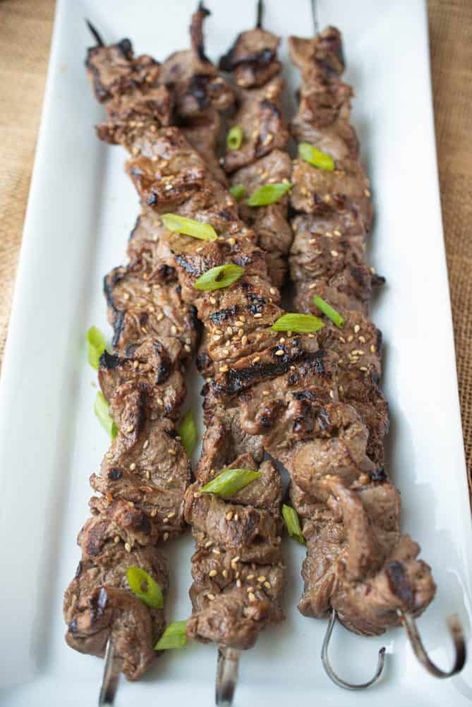 4 kabobs of Korean marinated beef on a rectangle plate topped with spring onion and sesame seeds