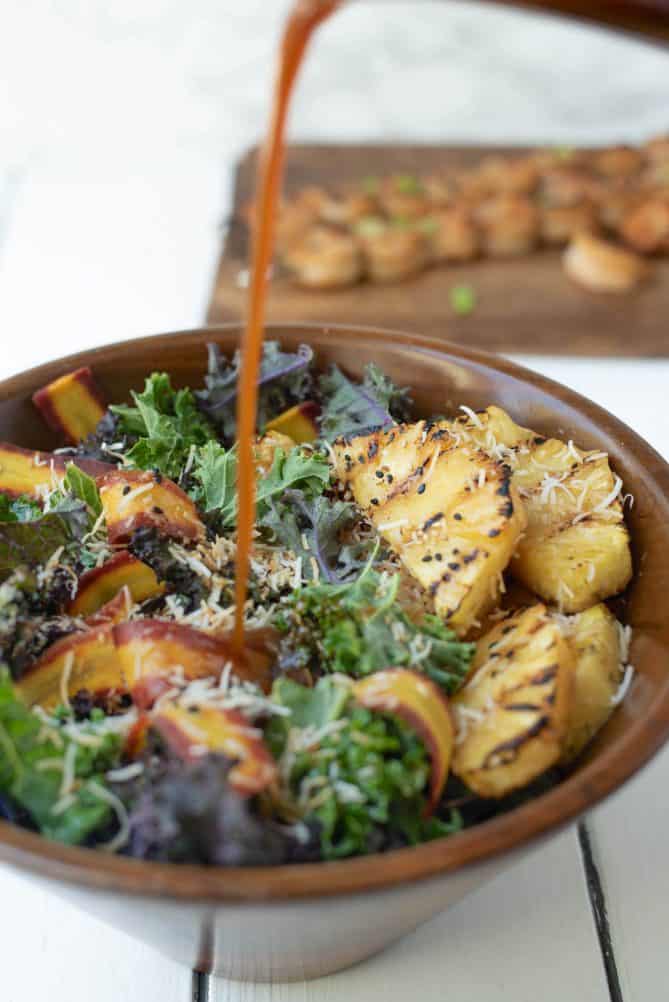 Pouring soy sesame dressing over a bowl of grilled pineapple coconut kale salad