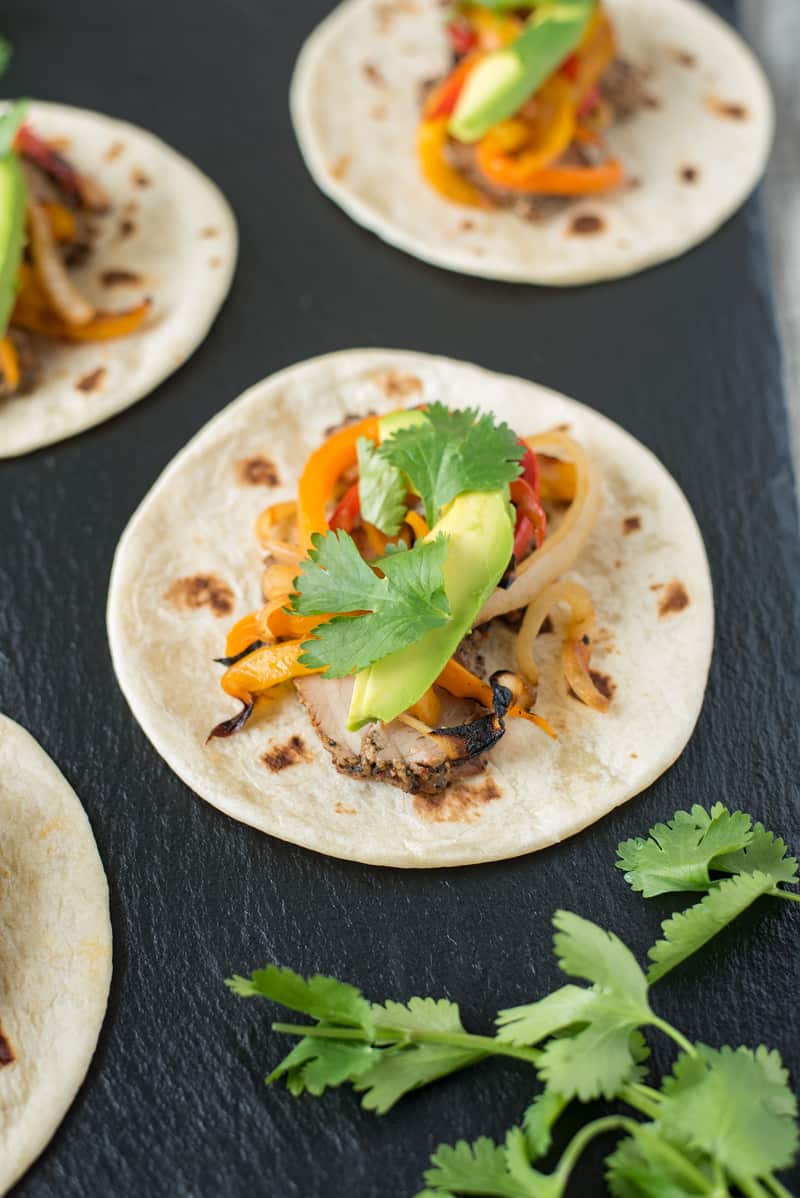 Grilled pork tenderloin fajitas are a quick and easy way to get a summer meal served in under 30 minutes. Marinated pork tenderloin is grilled along with onions and peppers. The pork is sliced then all are served on warm tortillas garnished with fresh avocado and cilantro.