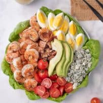 A large platter with lettuce leaves, grilled shrimp, tomato, bacon, egg, avocado and blue cheese