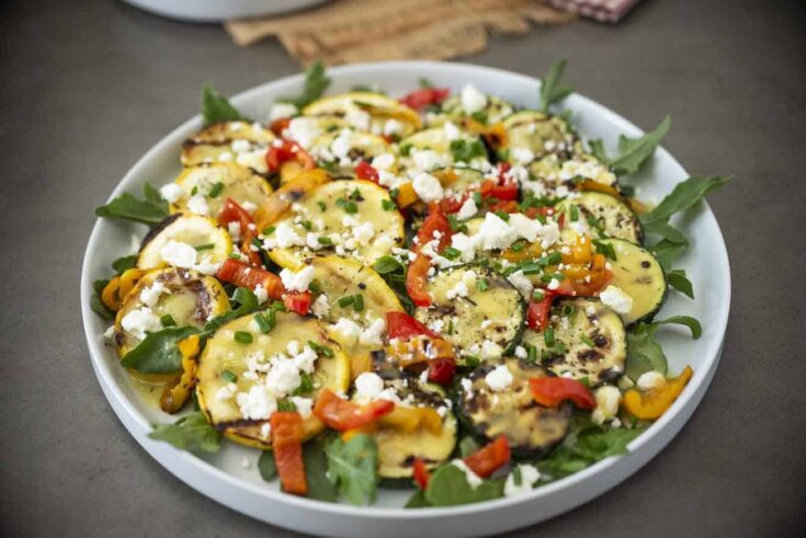 Zucchini and yellow squash on a white round platter with arugula peppers and feta