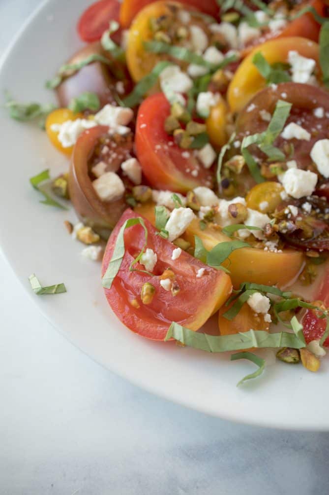 A closeup of a juicy, red and yellow heirloom tomato with chopped basil, feta and pistachios