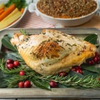 A full roast turkey dinner with a turkey breast on a serving tray with carrots in a white bowl, sweet potato casserole and a small container of gravy