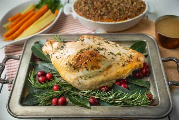 A full roast turkey dinner with a turkey breast on a serving tray with carrots in a white bowl, sweet potato casserole and a small container of gravy