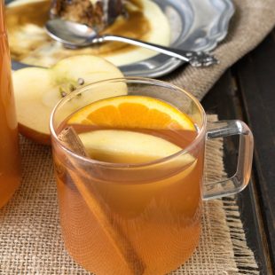 A glass mug of apple cider garnished with an apple wedge, orange slice and cinnamon stick