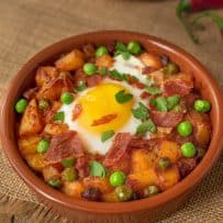 A terracotta bowl filled with potatoes, peas and chorizo in a tomat sauce topped with an egg