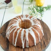 A whole bundt cake drizzled with frosting on a round, wood cake stand with lemons and daisies in the background