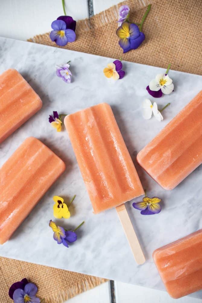 5 peach popsicles laying down on a marble board surrounded by edible purple, yellow and white pansy flowers