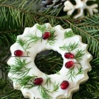 A cookie that looks like a Christmas wreath decorated with white frosting, green leaves and pomegranate seeds on fern