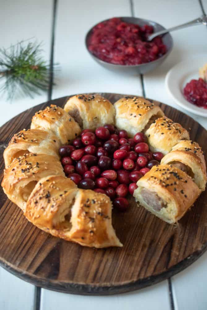 A puff pastry sausage roll wreath on a round board with 1 missing with fresh cranberries and a cranberry apple sauce in the background