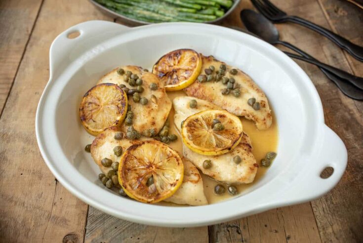 A white round dish of chicken piccata with lemon slices and capers.