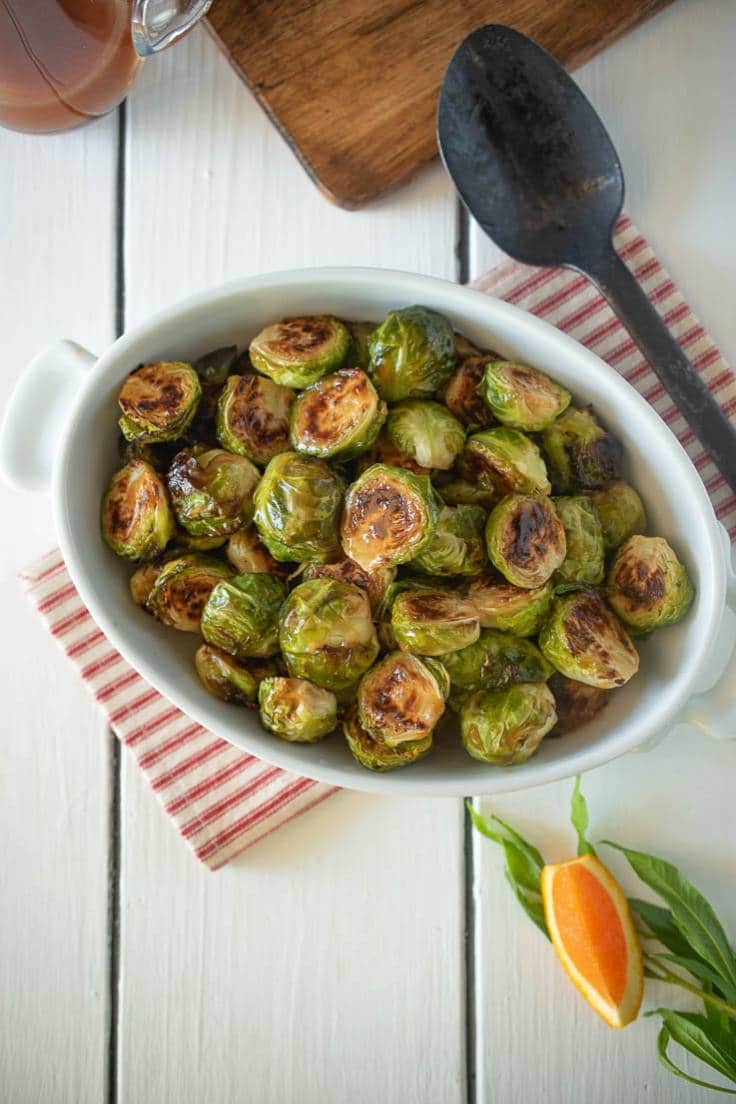 An overhead image of an oval bowl filled with roasted Brussels sprouts on a striped cloth with a serving spoon