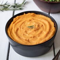 Vibrant orange puree in a black bowl garnished with fresh rosemary with a serving spoon