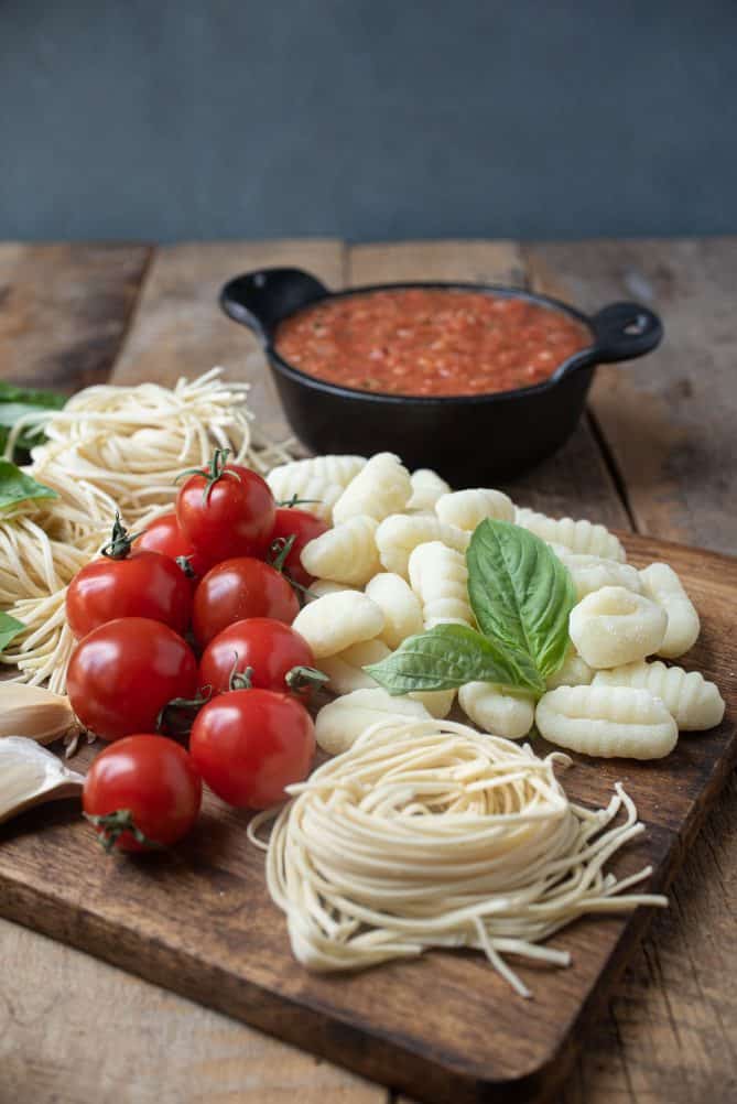 Fresh linguine, gnocchi, tomatoes and fresh basil on a board with tomato sauce in background