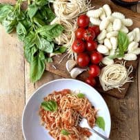 A display of fresh basil leaves, pasta and tomatoes viewed from overhead with a bowl of pasta and sauce