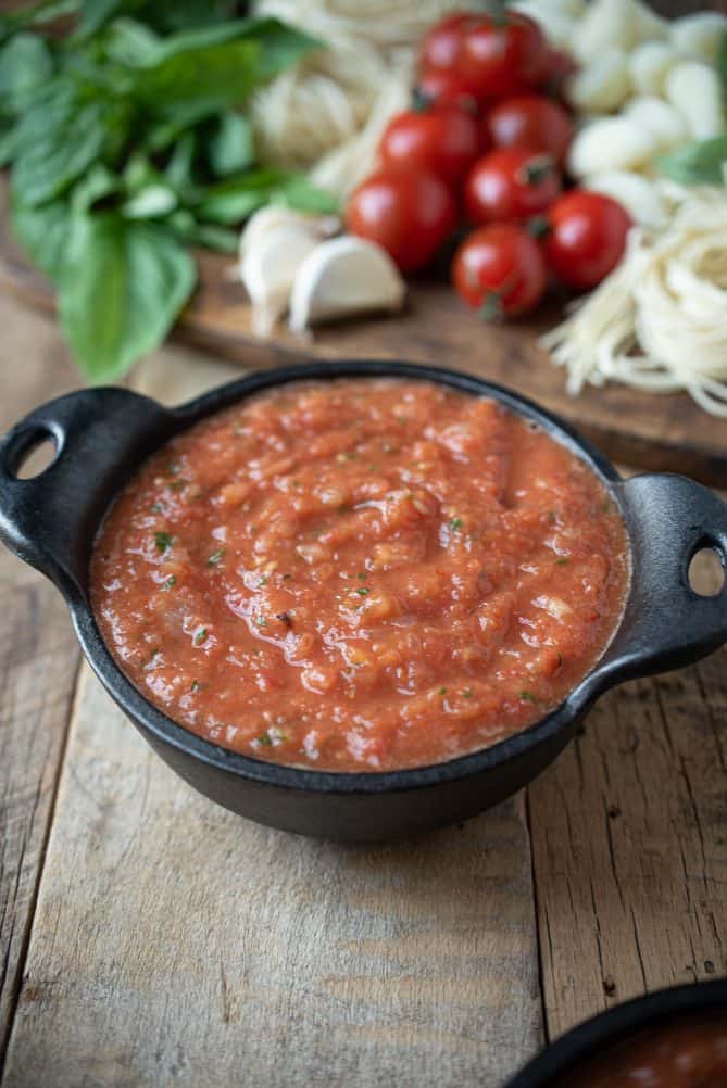Roasted tomato sauce in a black cast iron bowl with tomatoes, garlic, pasta and basil in the background