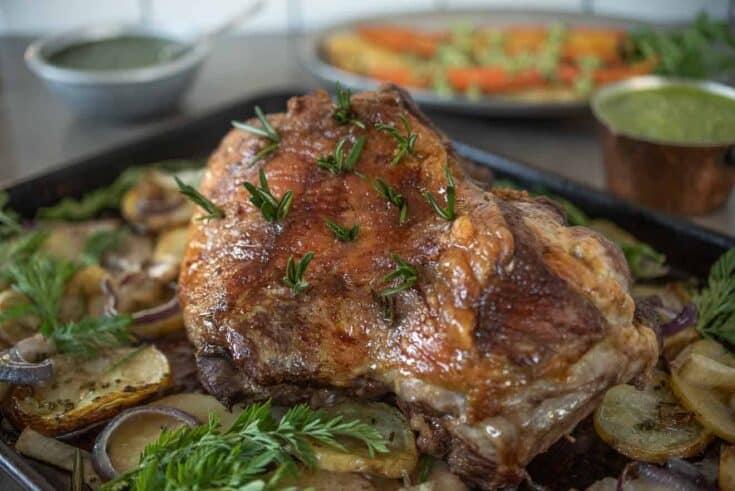 A perfectly browned, roasted leg of lamb in a roasting pan surrounded by vegetables