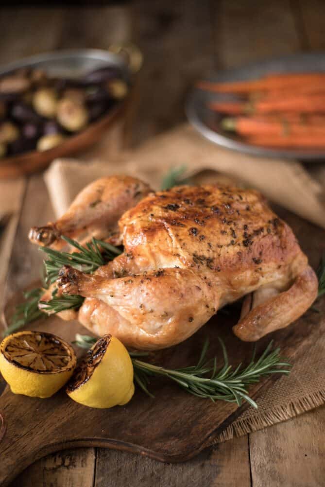 A browned, roast chicken in a cutting board with fresh herbs