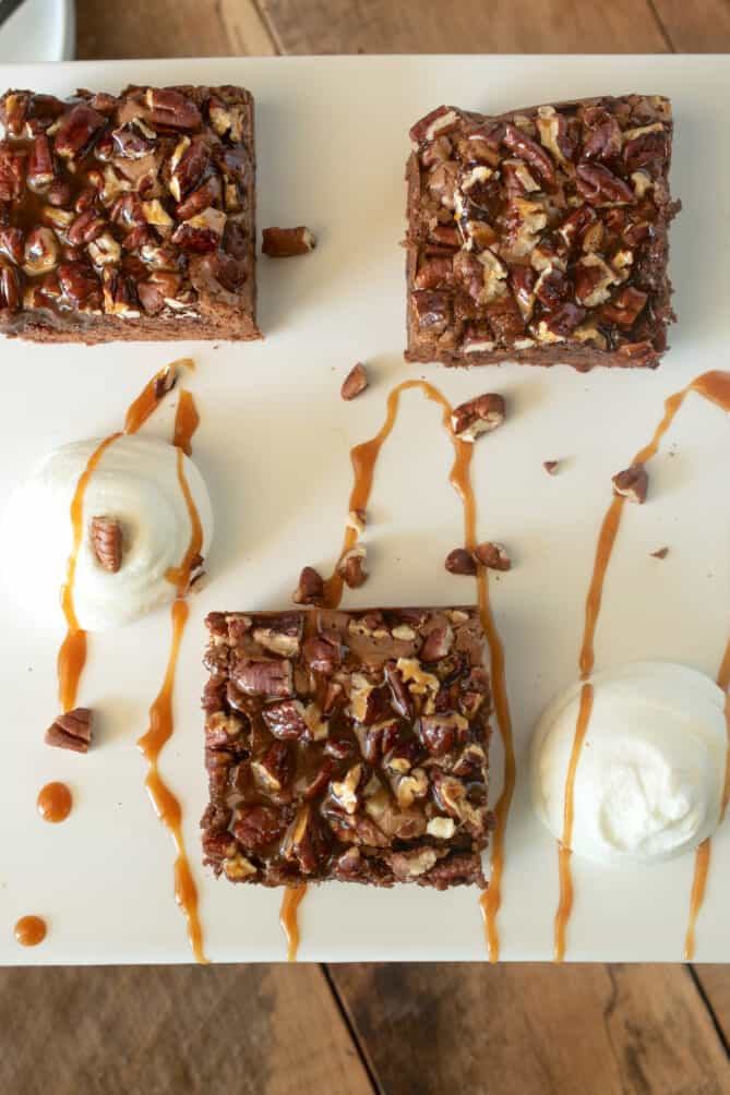 An overhead image of brownie slices on a white platter with whipped cream and caramel sauce