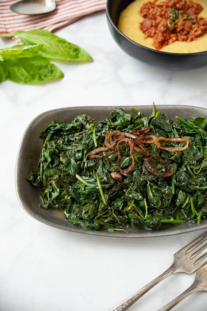 A pewter bowl filled with sauteed spinach topped with crispy shallots