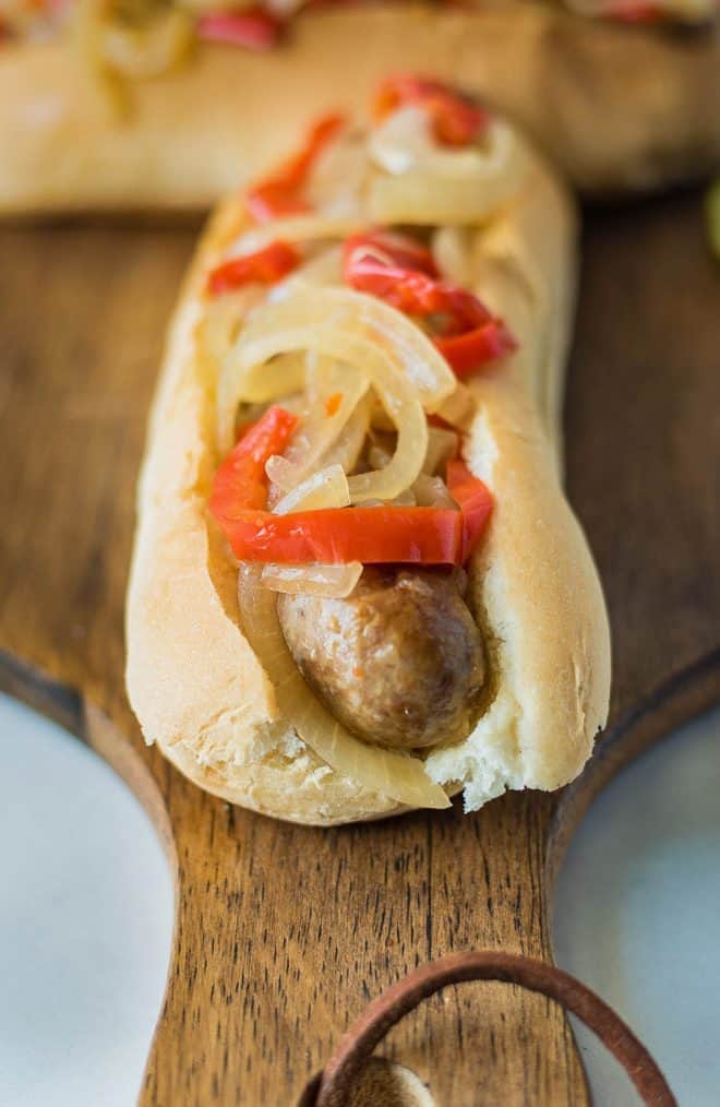 A closeup of a slow cooker beer bratwurst on a bun topped with onions and peppers
