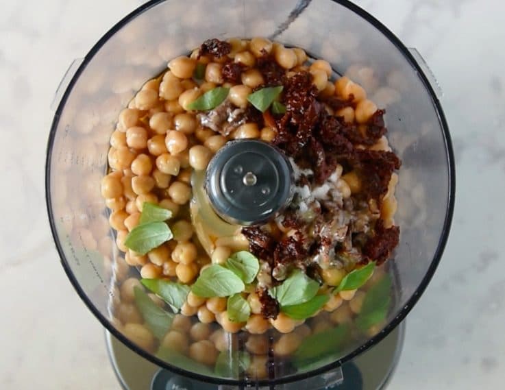 A food processor with chickpeas, sundried tomatoes, tahini, salt and pepper ready to blend into hummus