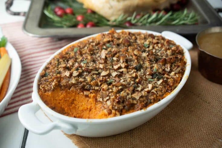 A white oval casserole dish filled with sweet potato casserole with pecan topping with a turkey in the background