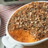 A closeup of a sweet potato casserole with a scoop taken out showing the chopped pecan topping flecked with fresh sage