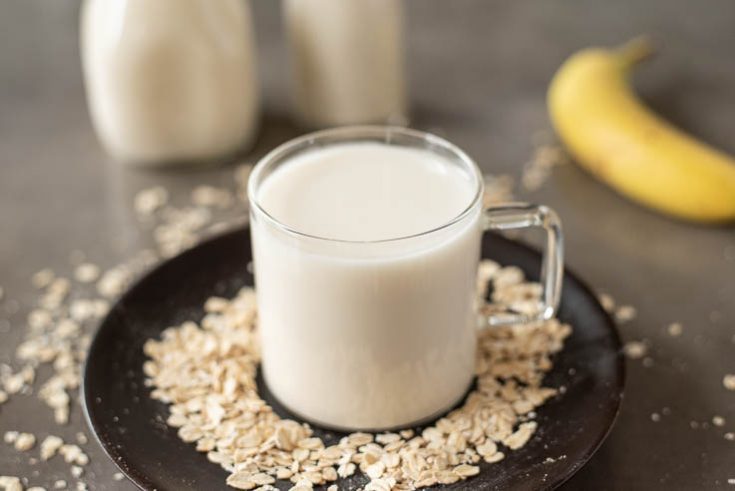Oat milk in a glass mug on a dark plate with bottles of oat milk and a banana